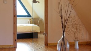 Un dormitorio con cama, una silla, un jarrón con ramas y suelo de madera.