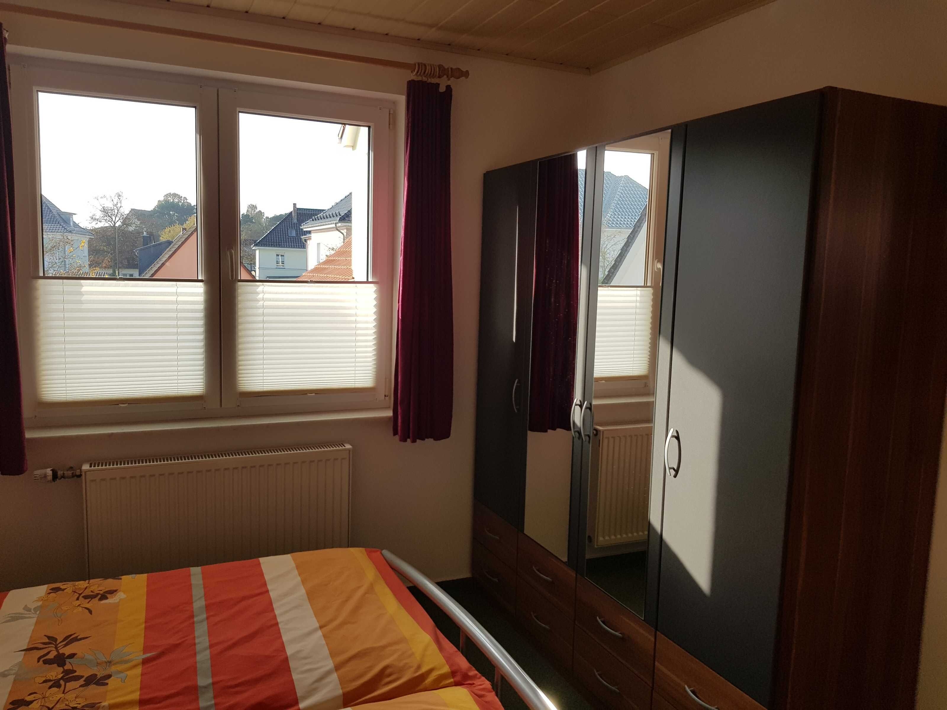 Ein Schlafzimmer mit Bett, Schrank, Heizkörper und einem Fenster mit Vorhängen.