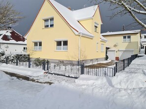 Une maison jaune à deux étages, dont le toit est recouvert de neige, et qui est entourée d’une clôture en métal noir.