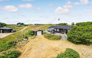 Main house with adjacent structure and outdoor seating area, set amidst grassy dunes and shrubbery.