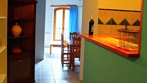 Een smalle keuken met een houten kast, een eettafel met stoelen en een aanrecht met een fles en mand.
