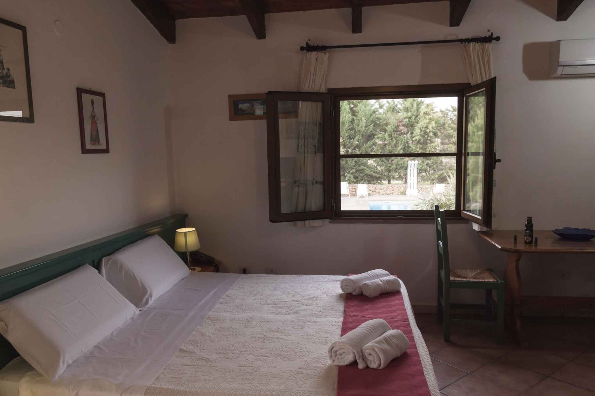 Habitació d'hotel amb llit, finestra amb cortines, taula i cadira, i una làmpada.