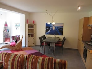 A cozy living room with a sofa, a dining table with chairs, a bookshelf, and a painting on the wall.