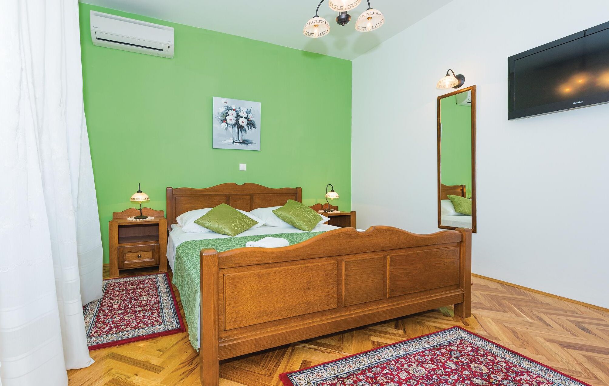 Un dormitorio con una cama de madera, paredes verdes, un televisor montado y una alfombra roja con estampado.