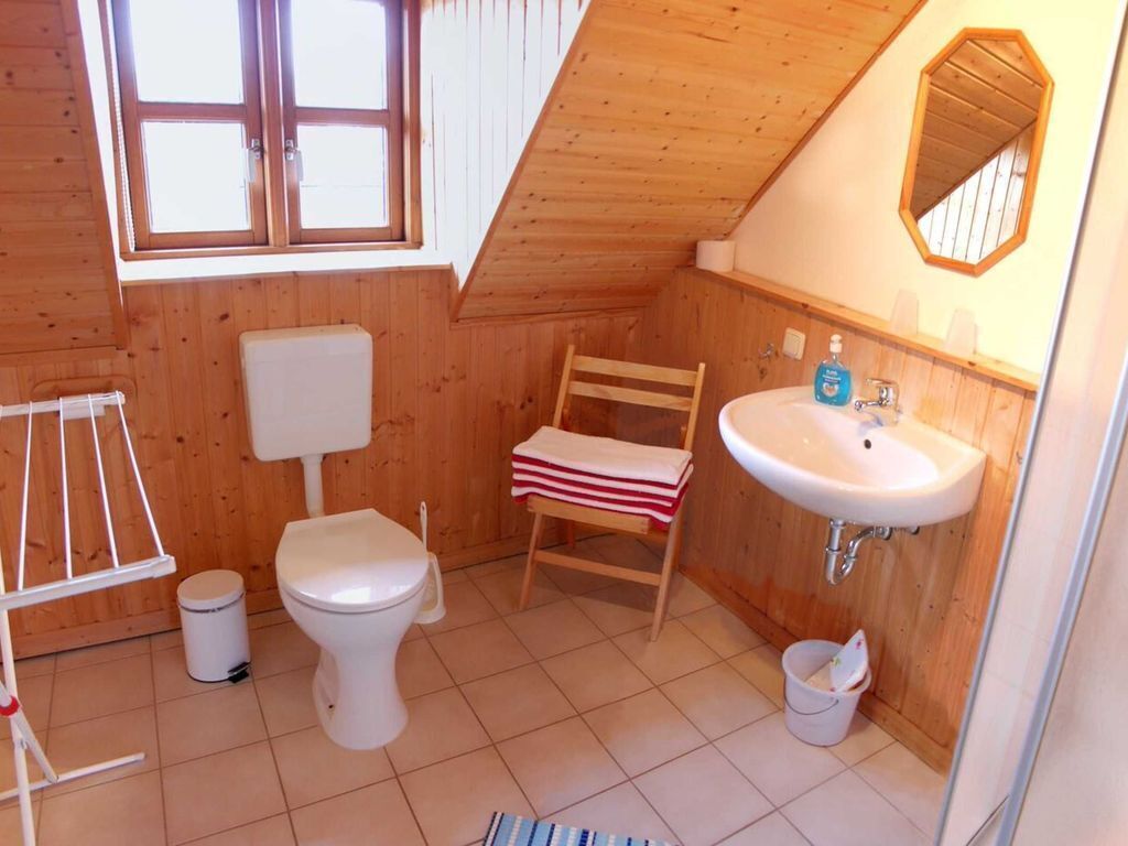 Une petite salle de bain aux murs lambrissés, équipée de toilettes, d’un lavabo, d’un miroir et d’une fenêtre.