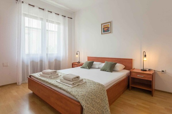 Una cama bien hecha con cabecera de madera, dos mesitas de noche con lámparas y una ventana con cortinas traslúcidas.
