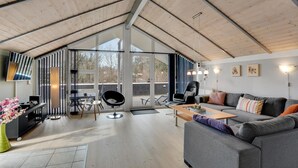 En rummelig opholdsstue med en stor grå sofa, et sofabord og en sort lænestol. Et tv er monteret på væggen, og et vindue giver naturligt lys.