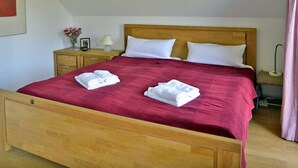 Un lit avec une tête de lit en bois, deux oreillers blancs et une couverture rouge. On y trouve une table de chevet en bois avec une lampe et un vase rempli de fleurs.