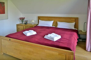 Ein Bett mit Holz-Kopfteil, zwei weiße Kissen und eine rote Tagesdecke. Ein Holznachtschränkchen mit Lampe und Blumen in einer Vase.