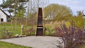 Un monument haut, étroit et de couleur sombre, orné d’inscriptions, entouré d’une zone pavée avec de l’herbe et des arbustes.