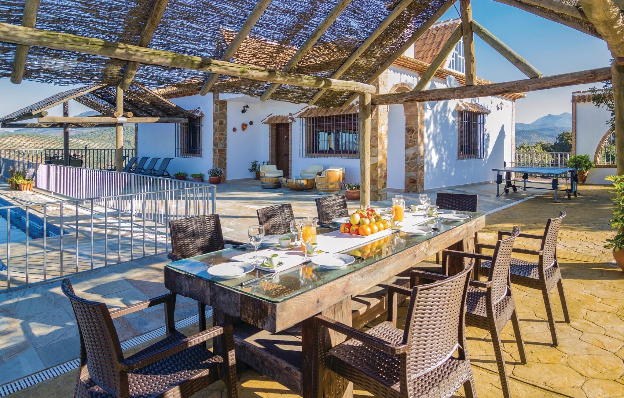 Uma área de refeições ao ar livre com uma mesa comprida, cadeiras de vime e uma piscina ao fundo.
