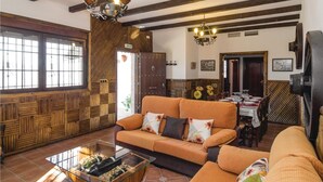 A cozy living room with a wooden wall, a sofa, a coffee table with bottles, and a dining area with a table set for four. - Beautiful home in Iznájar (Iznájar)
