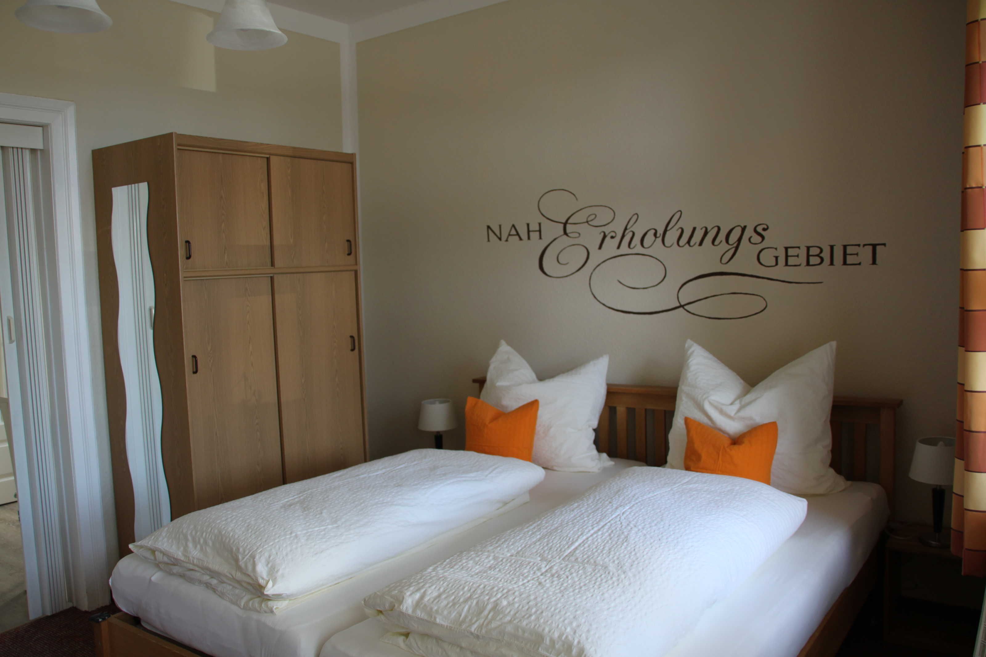 A hotel room with two beds, white bedding, and orange pillows. A wooden wardrobe is visible, and the wall has a sign that reads "NAH Erholungs GEBIET".