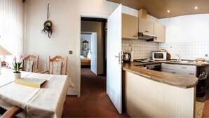 A cozy, well-lit room with a dining table, chairs, a lamp, and a view into a kitchen area with a microwave and sink. - Apartment E - Apartments Hasenpatt (Bad Sachsa)
