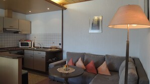 A compact living space with a kitchenette, a sofa with cushions, a small round table, and a floor lamp. - Apartment E - Apartments Hasenpatt (Bad Sachsa)