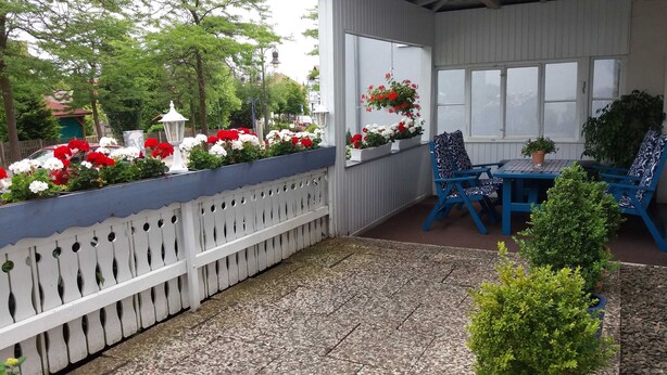A covered patio with a white picket fence, a table and chairs, and hanging flower baskets. - Apartment E - Apartments Hasenpatt (Bad Sachsa)