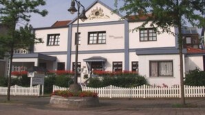 A two-story building with a white facade, a red roof, and a small porch area. There is a white picket fence in front of the building. - Apartment E - Apartments Hasenpatt (Bad Sachsa)