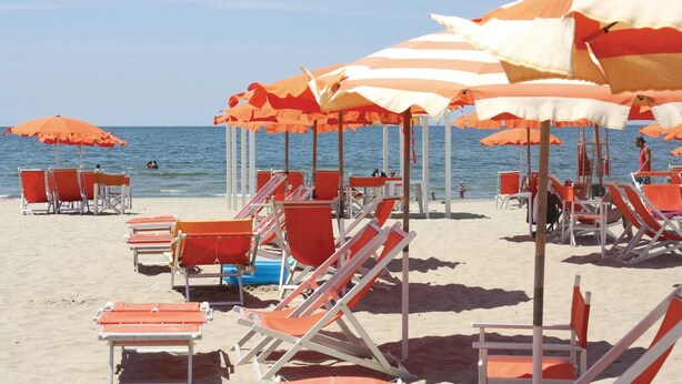 Una spiaggia con file di ombrelloni arancioni e bianchi e lettini rossi.