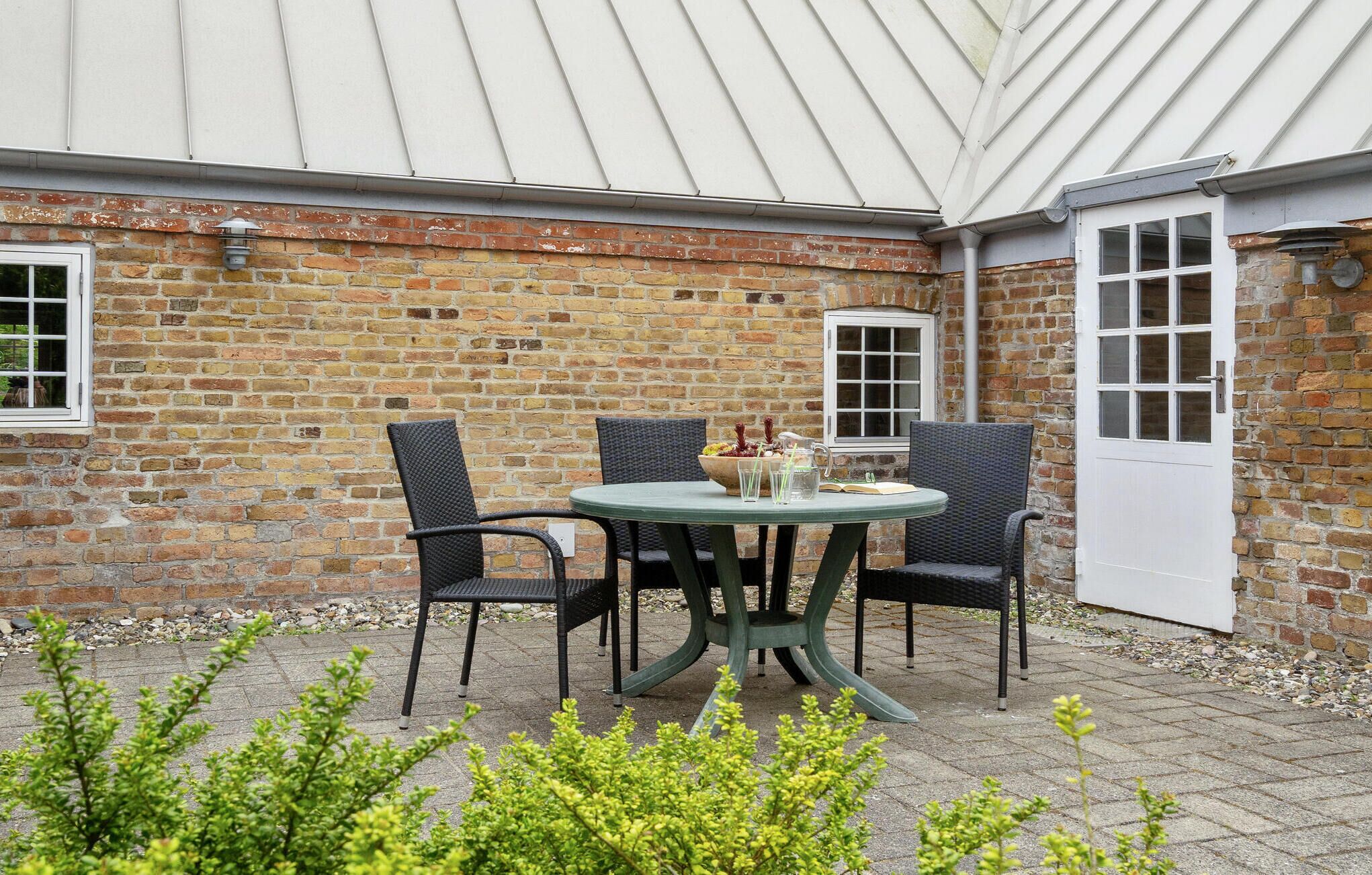Une terrasse extérieure avec une table ronde et quatre chaises, un mur en briques et une porte blanche.