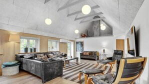A spacious living area with a large sofa, a coffee table, and a wooden floor. There are two chairs and a small table with a lamp. The room has a high ceiling with exposed beams and hanging lights. - Stunning home in Ulfborg with sauna (Ulfborg)