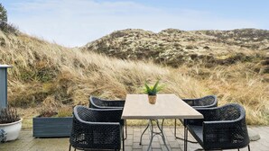 Eine Terrasse im Freien mit Tisch und vier Korbstühlen, mit Blick auf einen grasbewachsenen Hügel unter blauem Himmel.