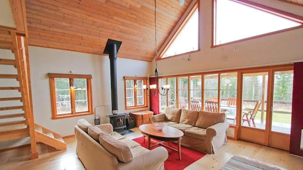 A spacious living room with a high ceiling, wooden beams, a fireplace, a staircase, and large windows offering a view of the outdoors. - Amazing home in Annerstad with sauna (Annerstad)
