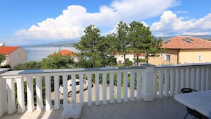 Ein Balkon mit weißer Reling, einem Tisch und Stühlen. Im Hintergrund sind Häuser mit roten Dächern, Bäume und ein Gewässer zu sehen.
