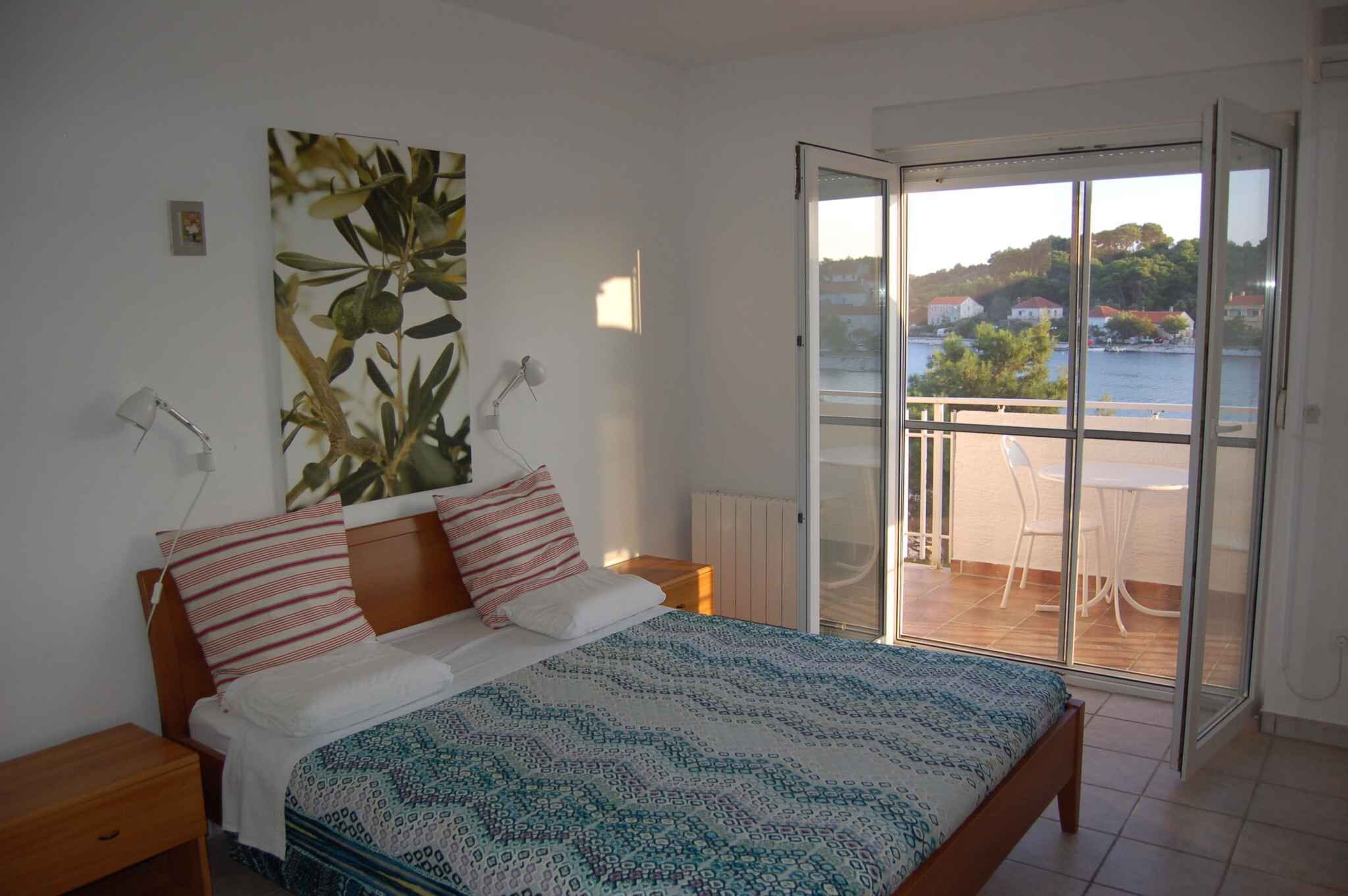 A bedroom with a bed, a nightstand, a wall-mounted lamp, and a sliding glass door leading to a balcony with a view of the sea and buildings.