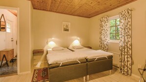 A bedroom with a wooden ceiling, a bed with two bedside tables, a window with floral curtains, and a small bedside lamp.