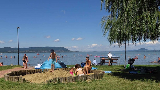 Ein Strand mit badenden und sonnenbadenden Personen, einem Zelt und einer Holzhütte am Ufer.