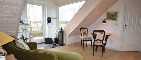 Un dormitorio tipo loft acogedor con un sofá verde, una silla de madera y una mesa de comedor. Hay un ventanal grande que proporciona luz natural y vistas al exterior.