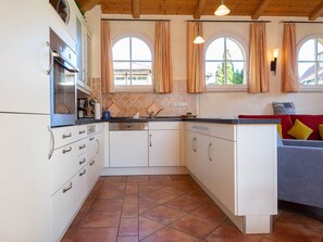 Una cocina compacta con muebles blancos, horno integrado y lavavajillas. Hay dos ventanas con cortinas, y un sofá con cojines en la zona de estar contigua.