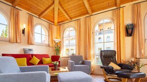 A spacious living room with a red sofa, grey armchairs, and a black leather chair. There are large windows with curtains, a wooden ceiling, and a tiled floor. - L-39 - Residence Laguna Whg L-39 .. (Kühlungsborn)