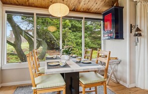 Un coin repas avec une table dressée pour quatre personnes, entourée de chaises en bois dotées de coussins. Une grande fenêtre offre une vue sur les arbres et une structure en bois à l’extérieur.