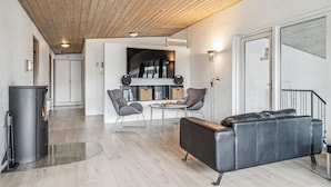 Ein modernes Wohnzimmer mit Kamin, einer Couch, einem Esstisch mit Stühlen sowie einer Küchenzeile mit Bücherregal und Pendelleuchte.