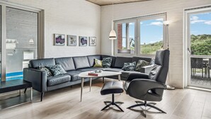 Ein modernes Wohnzimmer mit einem grauen Sofa, einem schwarzen Sessel, einem runden Couchtisch und einem Beistelltisch. An der Wand hängen vier gerahmte Kunstwerke, ein großes Fenster mit Blick ins Freie und eine Holzbalkendecke.