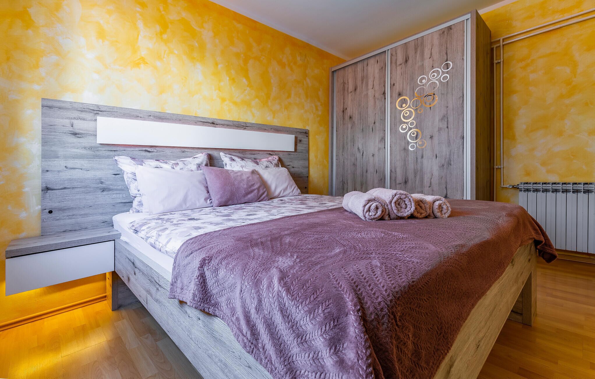 Quarto moderno com cabeceira de madeira, uma cama grande com manta roxa e um painel decorativo na parede.