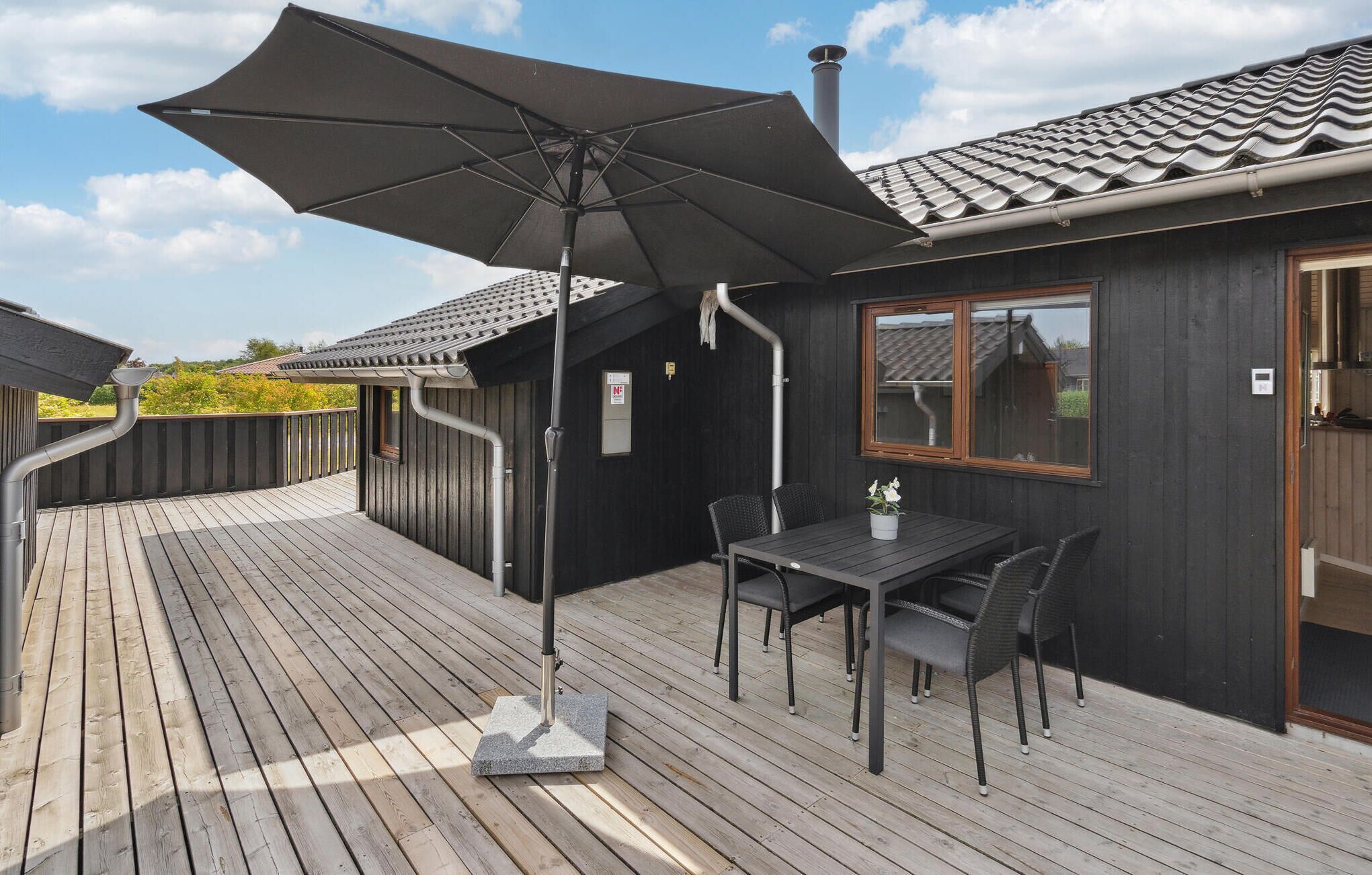 Une terrasse en bois avec une table et des chaises, un grand parasol et un bâtiment doté d’un toit en tuiles.