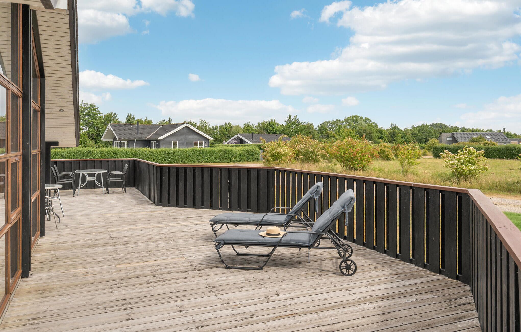Een houten terras met twee ligstoelen en een tafel, omgeven door een zwarte schutting, met uitzicht op een gazon met bomen en huizen op de achtergrond.