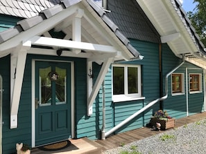 Una casa color turquesa con puerta principal blanca, un pequeño porche y una jardinera de madera con flores.