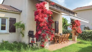 Ein Haus mit einer Terrasse, auf der ein Grill, Sitzmöbel und leuchtend rote, an den Wänden hochrankende Blumen zu finden sind.