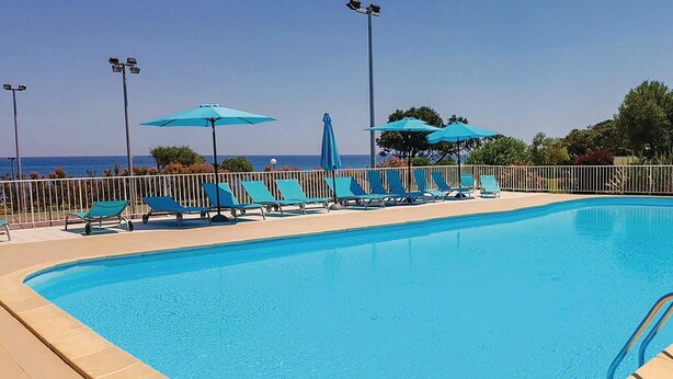 Ein Swimmingpool mit blauen Stühlen und Sonnenschirmen, umgeben von einem Zaun und mit Blick aufs Meer.