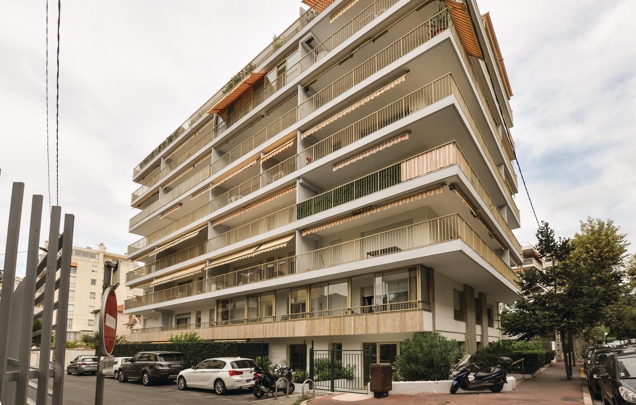 A multi-story residential building with balconies, a gated entrance, and parked cars.