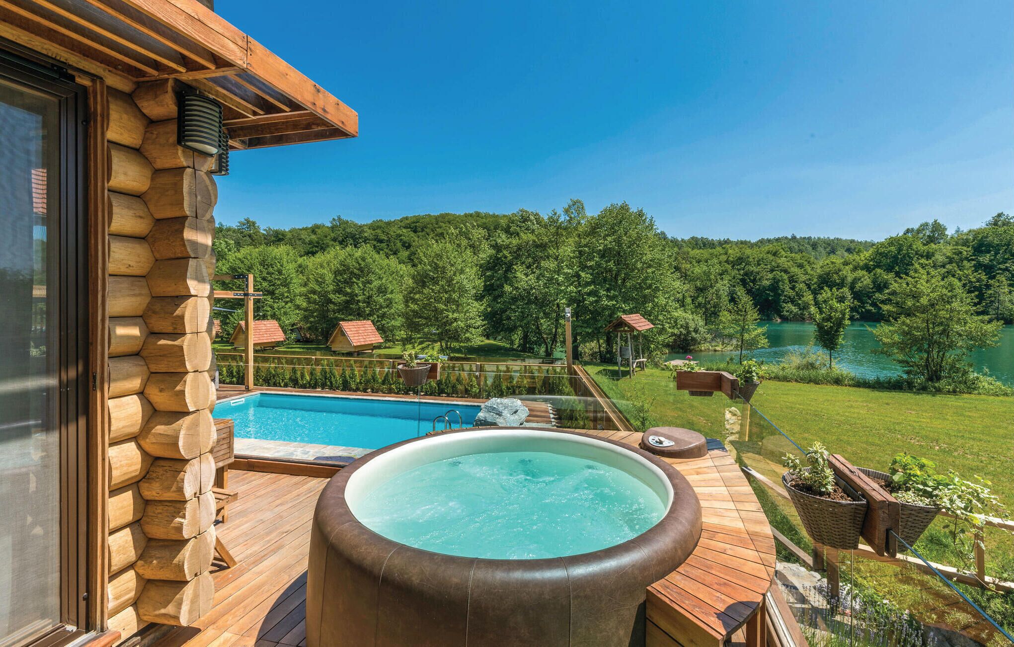 A wooden deck with a hot tub, a swimming pool, and a view of a lake surrounded by trees.