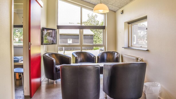 A room with a dining table and four chairs, a television mounted on the wall, and large windows providing a view of the outside.