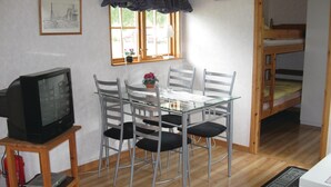 Quarto compacto com mesa de vidro, quatro cadeiras, televisão sobre suporte, cama beliche, relógio e quadro na parede.