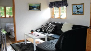 Sala de estar com sofá de couro preto, mesa de centro branca e duas janelas com cortinas. Há um quadro na parede e um vaso de planta no parapeito.