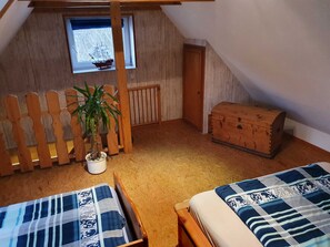 Ein Zimmer mit schrägen Wänden, einem Fenster mit einer Schiffmodell-Darstellung, einer hölzernen Truhe, einer Pflanze und zwei Betten mit gemusterten Bettbezügen.