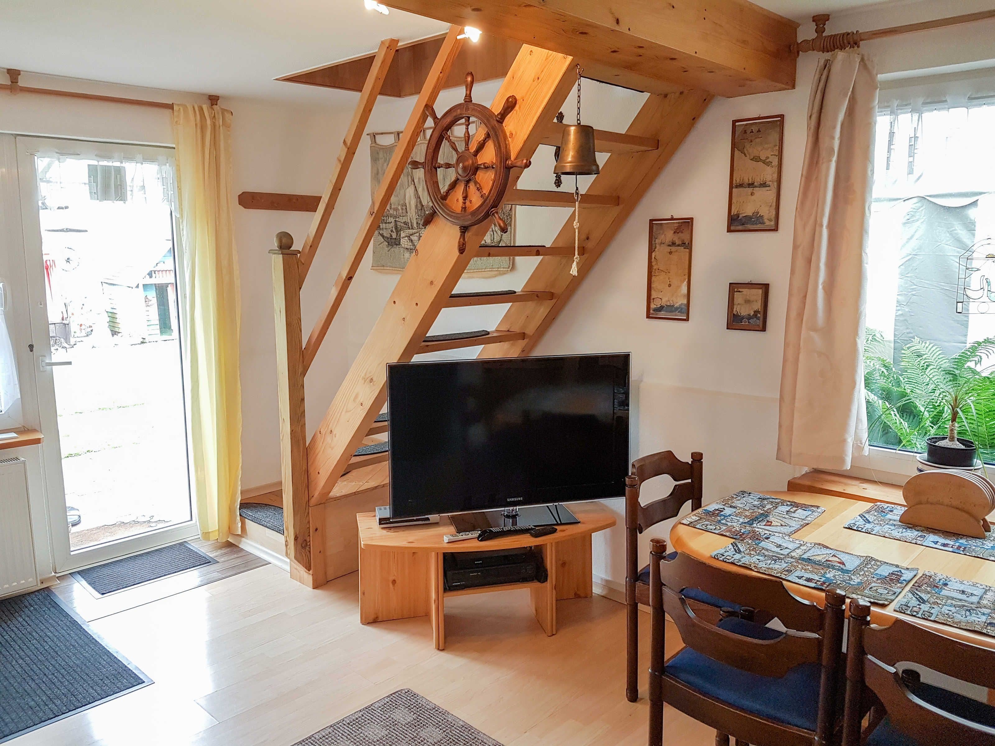Ein Zimmer mit einer Holztreppe, einem Fernseher auf einem Holztischchen, einem Esstisch mit Untersetzern und einem Fenster mit Blick auf Grünflächen.
