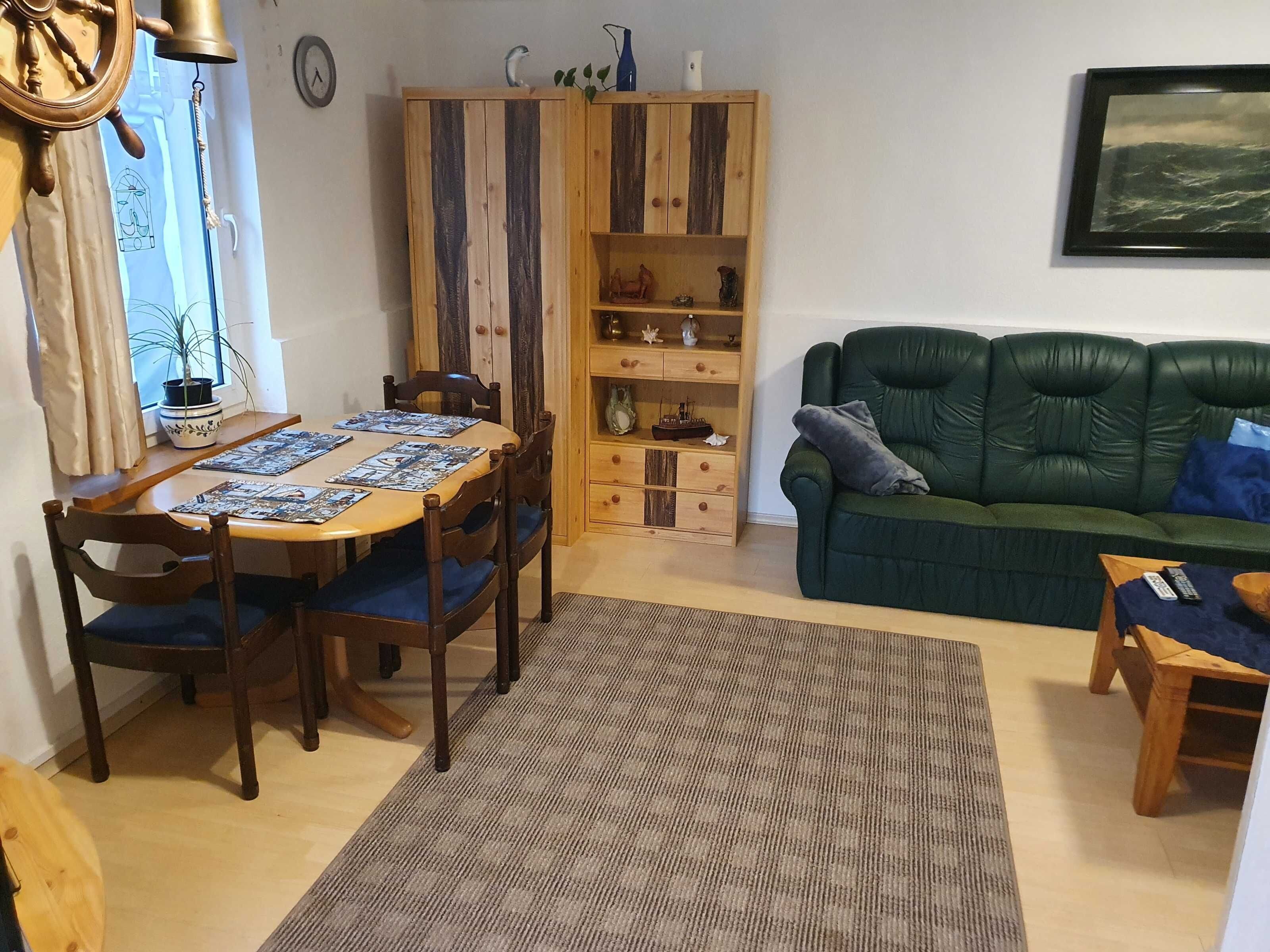 Ein Essbereich mit Holztisch und -stühlen, einem grünen Sofa, einer Holzkabine mit Regalen und einer Wanduhr.
